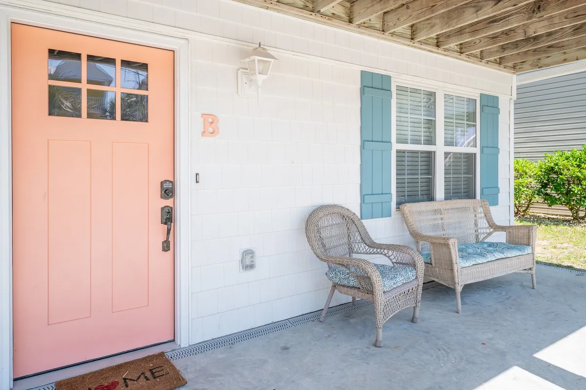 This inviting exterior shot showcases the home's coral front door, woven chairs, blue shutters, and a bright welcome mat. The setting suggests a welcoming environment that encourages guests to gather, relax, and enjoy outdoor moments together.
