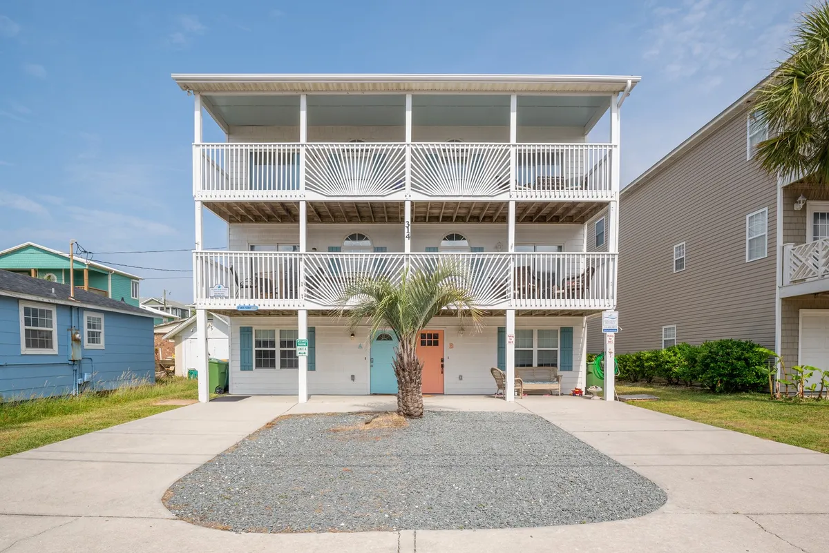 An exterior view of a two-story home featuring a spacious balcony, visible palm tree, colorful front door, and a gravel parking area suitable for multiple vehicles. The inviting scene hints at opportunities for outdoor gatherings and easy parking.