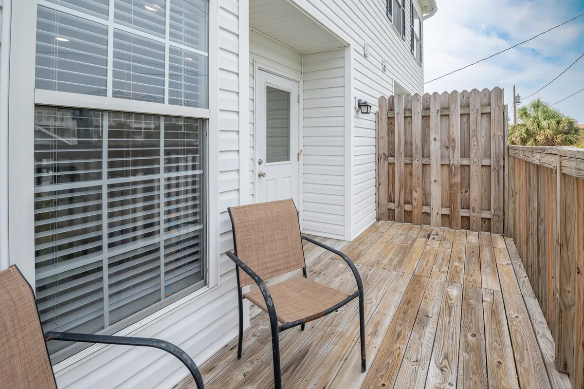 A covered wooden deck with two chairs, fencing, and a doorway leading inside, suggesting a seamless flow between indoor spaces and the outdoors. The deck looks out onto a tranquil setting, ideal for relaxing or lingering over views.