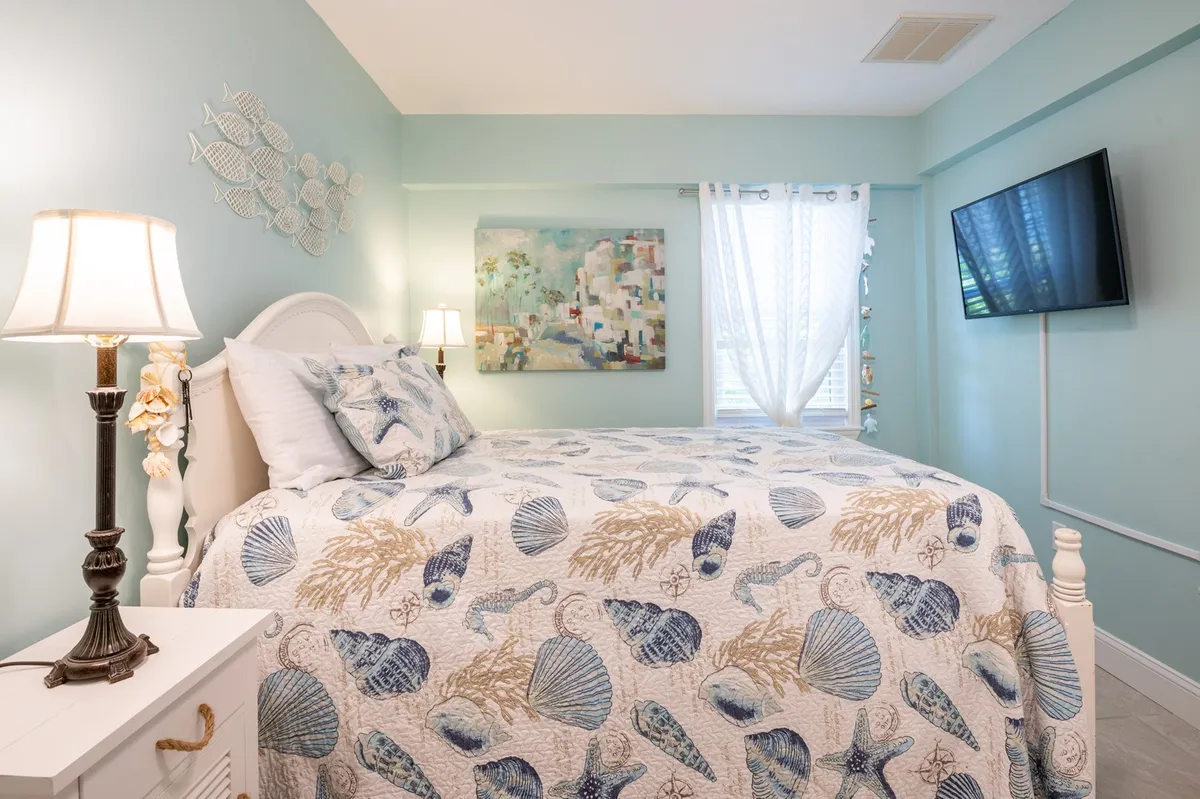 A serene bedroom with a bed featuring a seashell-patterned quilt, an abstract painting above the headboard, a lamp casting a soft glow, and a window draped with sheer curtains that filters natural light into the space. The overall decor emphasizes comfort and artistic inspiration.