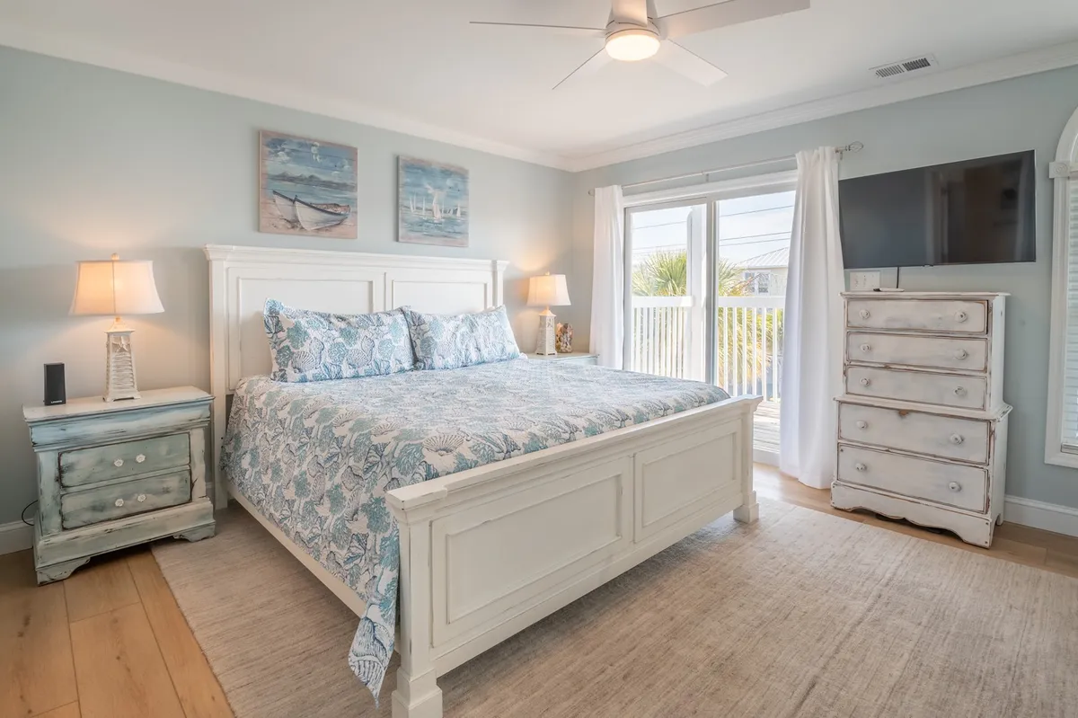 A cozy bedroom with a bed adorned in patterned bedding, a nightstand topped with a lamp, artwork on the wall, and a large window that offers an inviting view outside. The space combines natural light with a tranquil, homey ambiance.