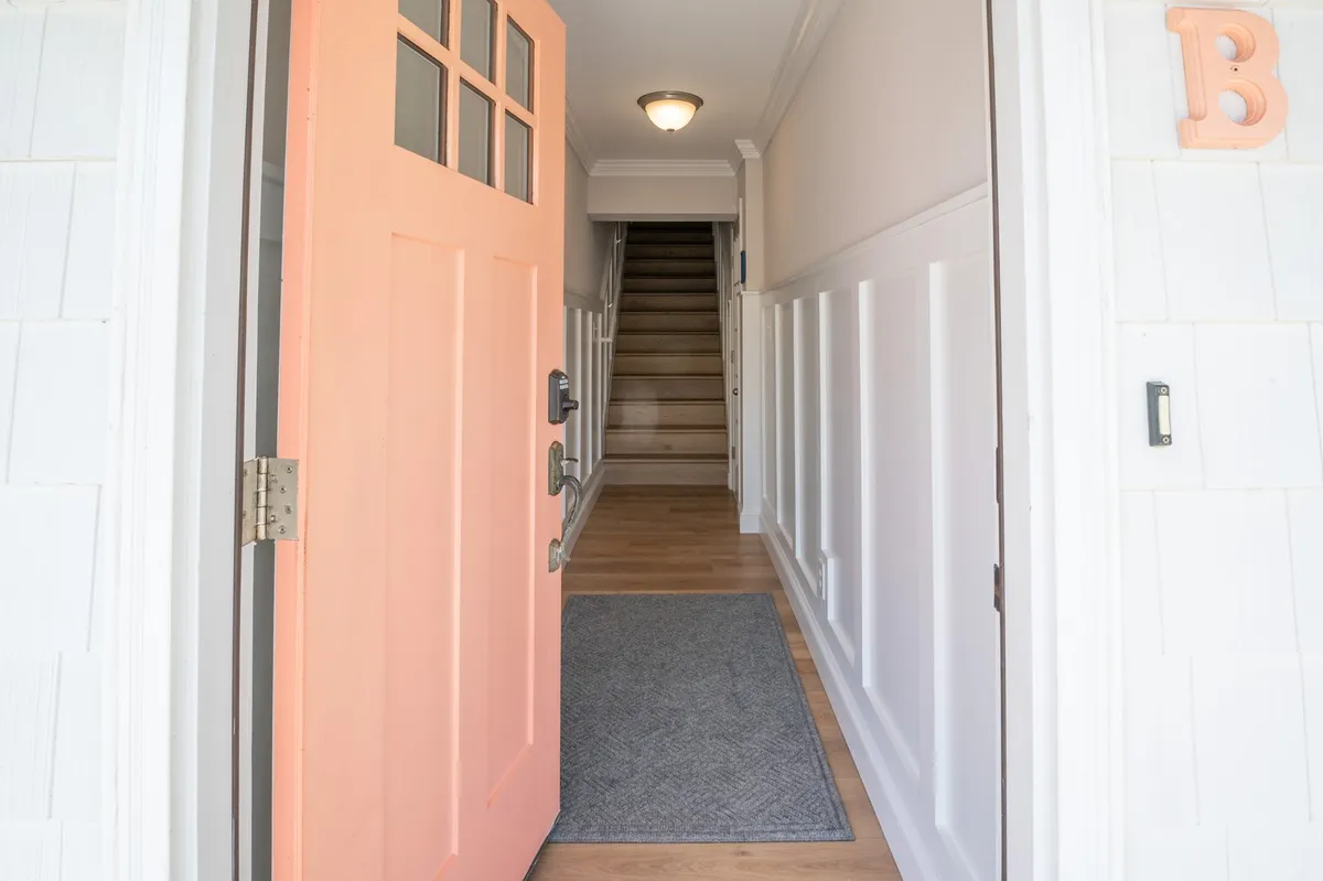 A bright, coral-colored front door opens to a staircase leading to the upper floors, with a textured area rug adding warmth. This image captures the moment of arrival, suggesting adventure and new experiences just beyond the threshold.