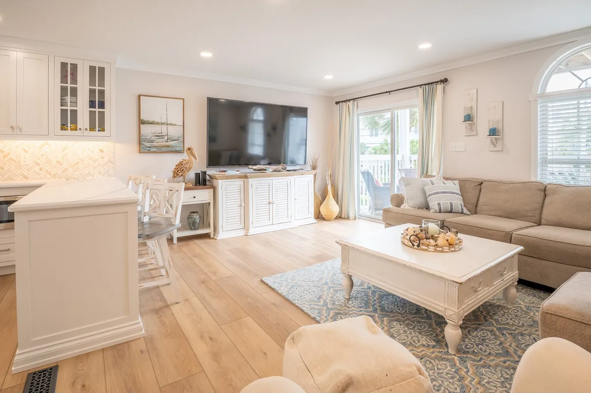 This image shows a welcoming open-concept living area with a plush sofa, coffee table, and a television visible in the background. The kitchen area and decorative art are also included in the frame, conveying a sense of space, comfort, and togetherness. Natural light streams in, brightening the communal layout.