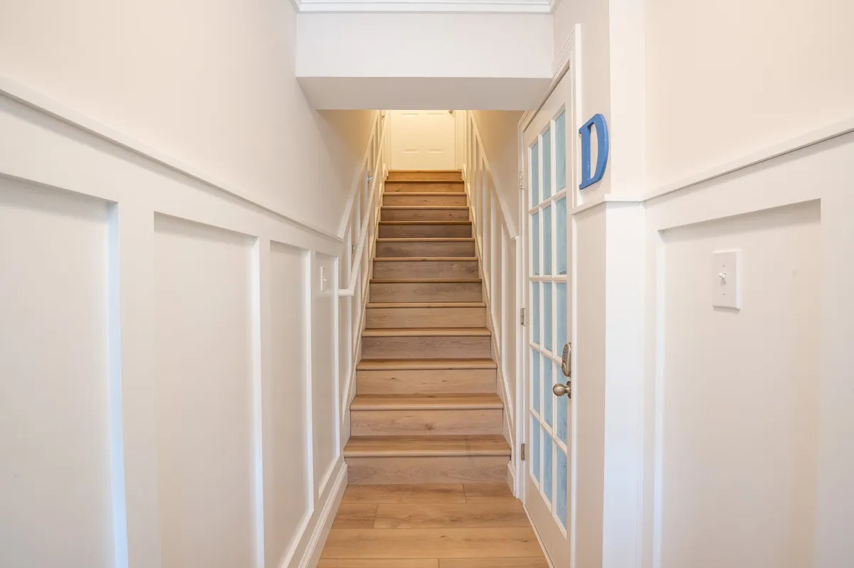 A welcoming interior hallway anchored by a stylish staircase and a glass door, with guests either returning home or arriving, suggesting movement between levels. Natural light and carefully chosen wall paneling give the entrance a bright, organized feel.
