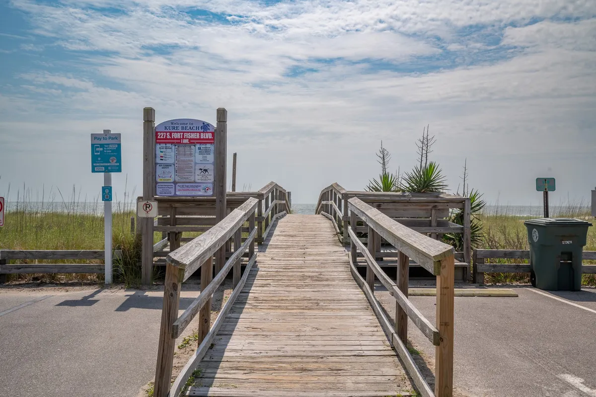 A wooden walkway bordered by lush green vegetation leads directly toward a 'Beach Access' sign, with parking and residential scenery hinting at the home's convenience to the beach. The perspective invites viewers to imagine the easy, flat path from the house to the ocean.