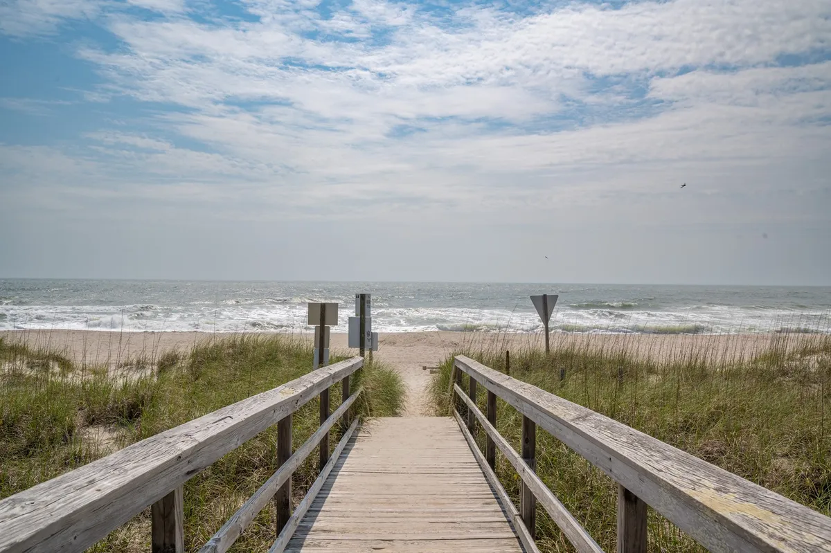 This image captures a scenic stretch of boardwalk leading over the dunes to the ocean, with blue sky overhead and gentle waves rolling in. The view suggests early morning or late afternoon, emphasizing the relaxing ambiance of the beach. Families and groups can be envisioned beachcombing, sunbathing, or simply enjoying the surroundings.