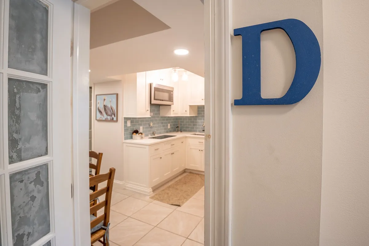 A bright kitchen area with kitchen cabinetry, a stylish backsplash, and a dining table set for family meals. The large blue letter 'D' adds personality and a sense of place. The space is warm and functional, ideal for both cooking and casual family dining.