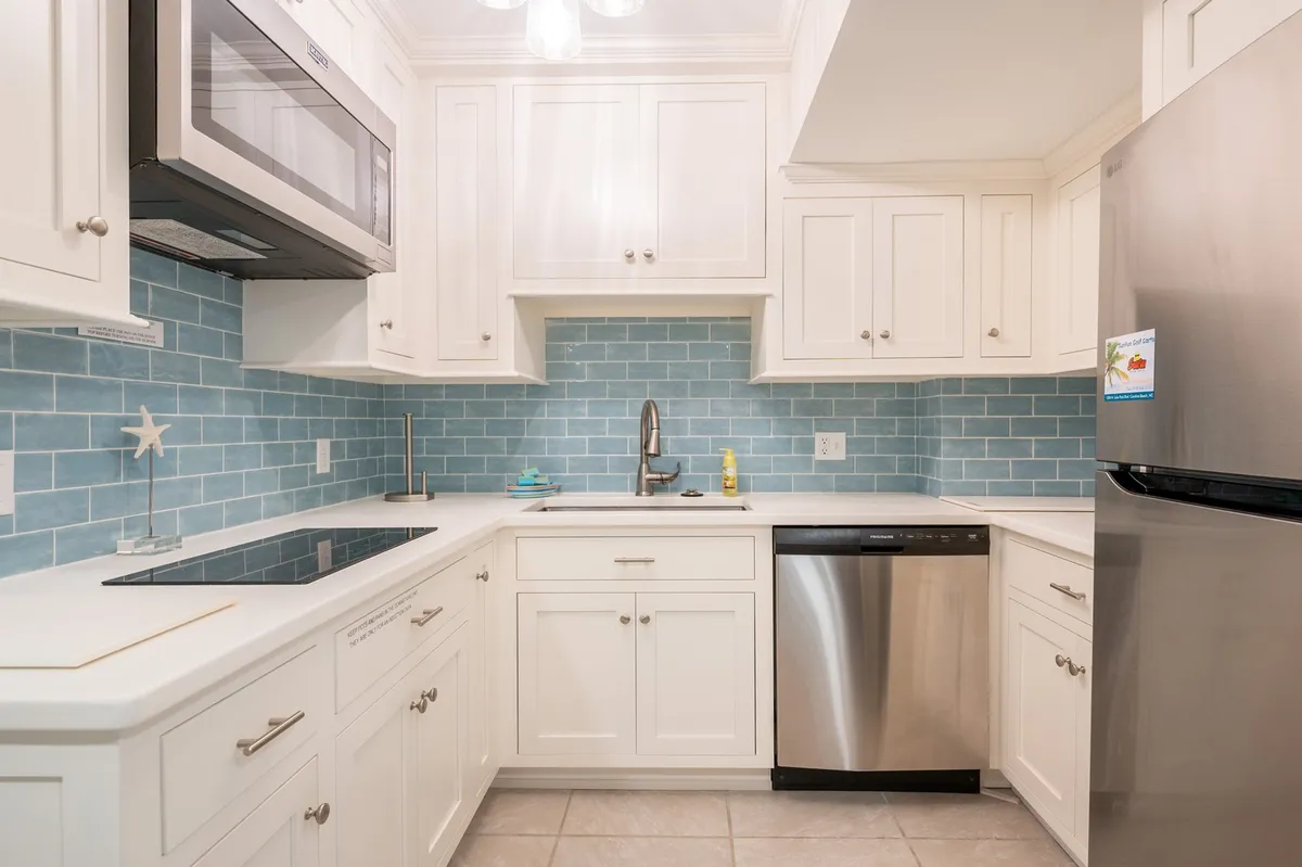 A newly remodeled kitchen featuring blue backsplash tiles, stainless steel appliances, white cabinetry, and clean countertops. The space is bright and organized, perfect for group cooking and socializing.