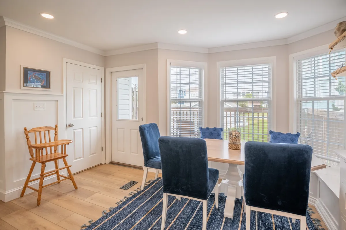 A spacious dining area with a wooden dining table surrounded by blue upholstered chairs and a wooden high chair, set in front of large windows that let in abundant natural light. The scene radiates warmth and readiness for shared meals and special occasions.