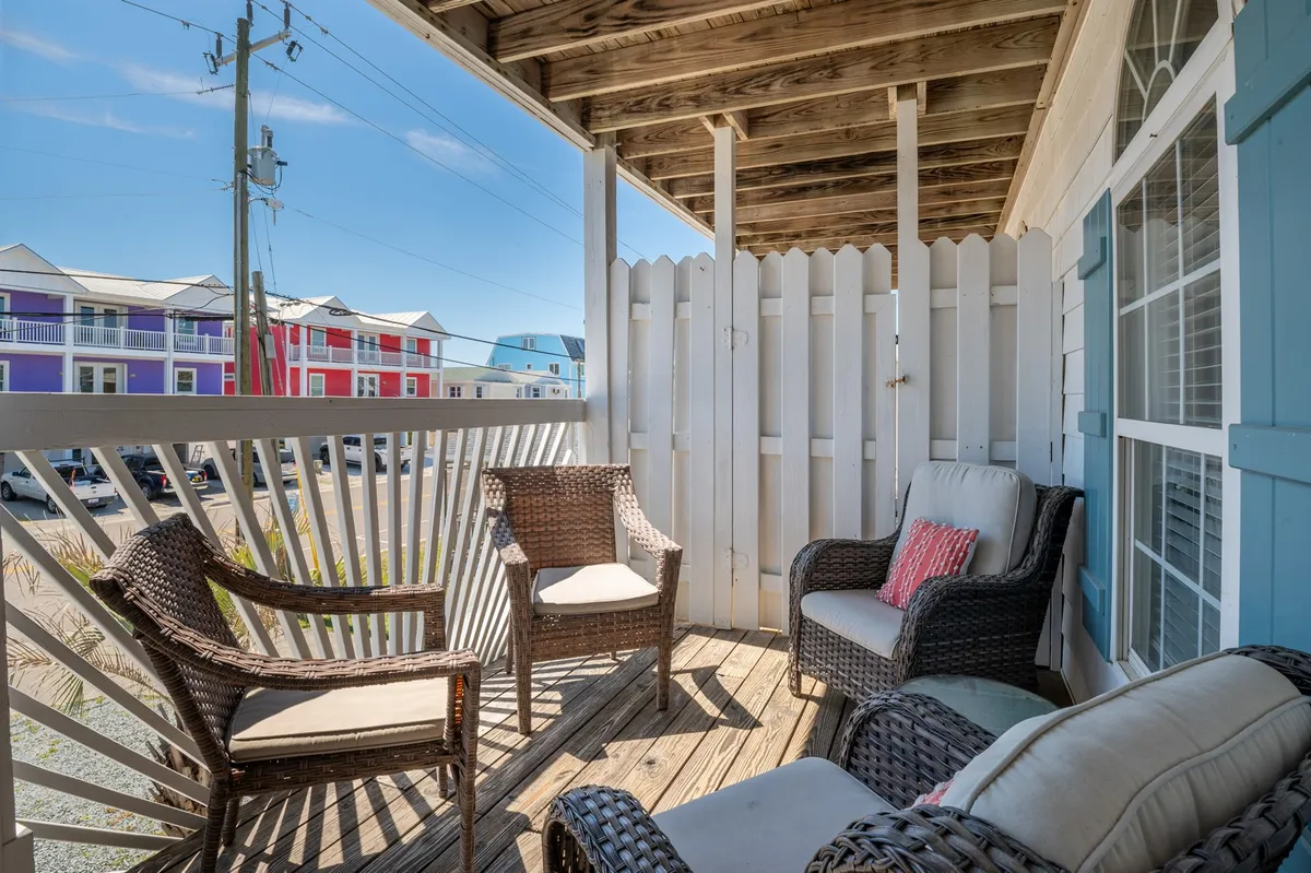 The image shows a bright, seaside wooden deck with comfortable wicker chairs and inviting blue accents, all framed by a classic white fence. The setting overlooks the ocean just across the street, inviting guests to relax, watch sunsets, or sip drinks while enjoying the tranquil coastal atmosphere.