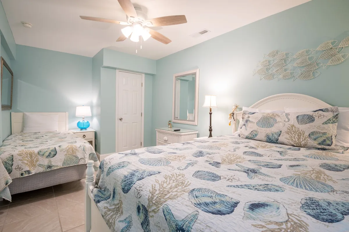 A light-filled, tranquil bedroom featuring two beds with seashell-patterned bedding, light blue walls, and a ceiling fan overhead. The morning sunlight streams through the window, creating a welcoming setting ideal for relaxation and shared family moments.