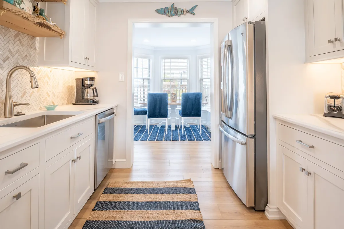 A compact yet functional kitchen setup with stainless steel appliances, white cabinetry, a chevron backsplash, and blue accent chairs. The striped rug and cozy seating area showcase a space ideal for quick snacks or quiet coffee breaks, reflecting a secondary kitchenette vibe.