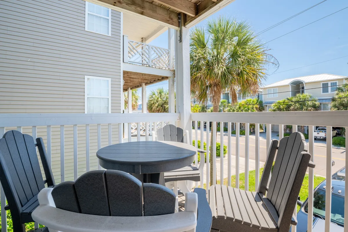 A relaxed exterior scene showcasing inviting outdoor seating under palm trees, with a clear view of the street and ample space to sit and enjoy a morning beverage. The porch area is positioned to take in sunrise views and casual outdoor moments.