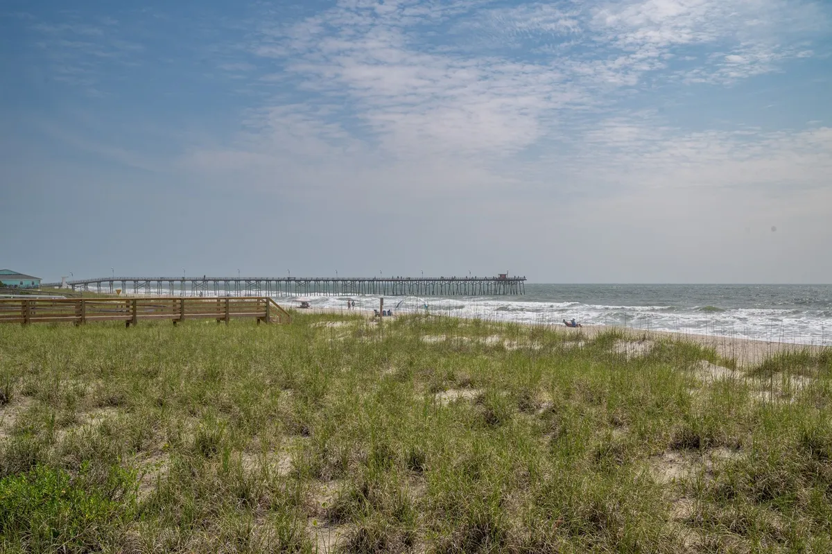 A wide view showcasing beach grass in the foreground, ocean waves rolling onto the sand, and the iconic Kure Beach Pier extending into the water under a bright blue sky. The scene encapsulates beachcombing, fishing, and families building sandcastles, perfectly representing the area's most beloved activities.